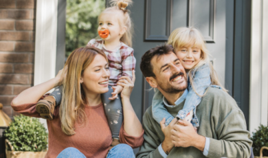 Couple sit on a porch stoop with two young children cuddling behind them and smiling at the camera.