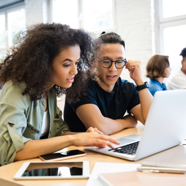 Young curly hair woman discussing with man in classroom. Classmates are learning through laptop. They are wearing casuals.