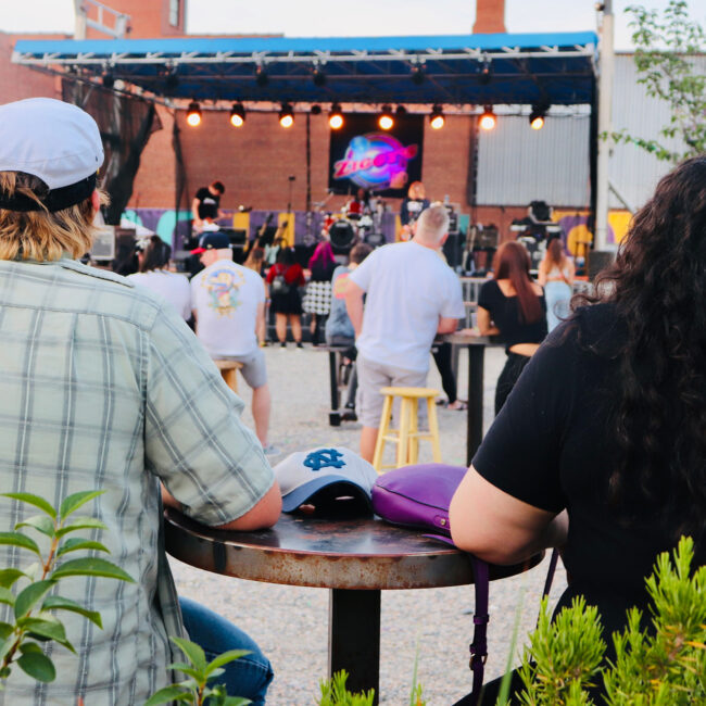 Two people sitting at a high-top table watching an outdoor live performance.